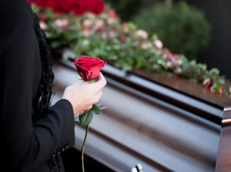 Woman holding a stem of rose while standing in front of a casket
